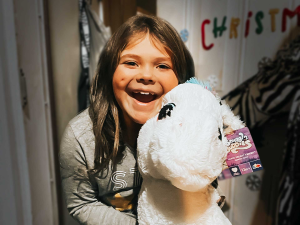 A child smiles while holding a large plush unicorn toy indoors. The word "Christmas" is partially visible on the wall behind, hinting at the festive excitement soon to be highlighted in the family newsletter.