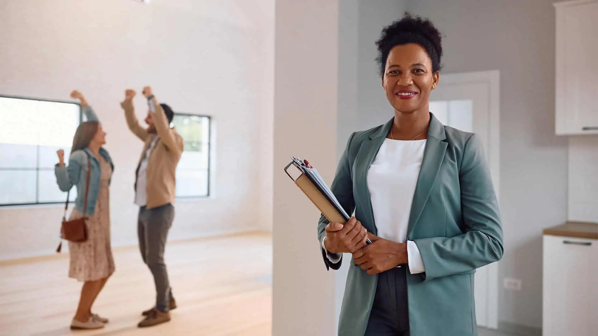 A real estate agent holding documents stands in the foreground, smiling. In the background, a happy couple celebrates in an empty room, envisioning a bright future thanks to seamless property, landlords property management