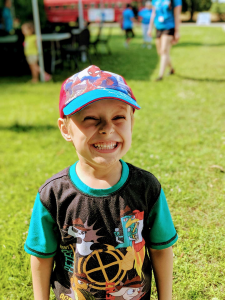 A smiling child wearing a superhero cap and printed shirt stands on grass in a park setting.
