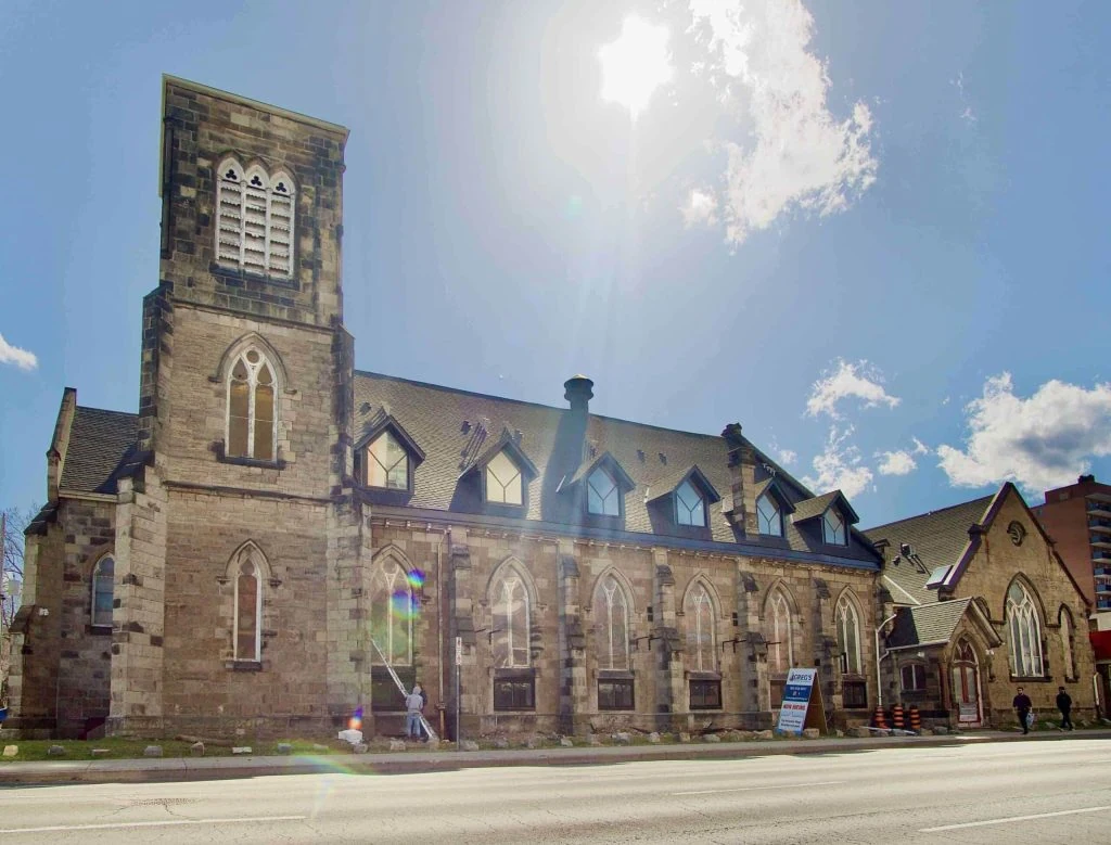 A historic stone church in Hamilton with a tall bell tower stands under a bright sun, casting lens flares. The sky is partly cloudy, and a few people walk by on the pavement, possibly admiring what could one day be an iconic Ontario rental property.