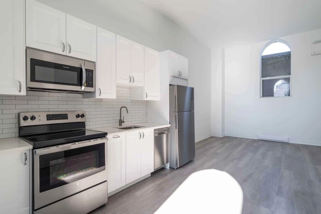 Located in Hamilton, this Ontario rental property features a modern kitchen with stainless steel appliances, including a microwave, stove, and refrigerator. Enjoy the elegance of white cabinets and a subway tile backsplash. The space is complemented by hardwood floors and an arched window.