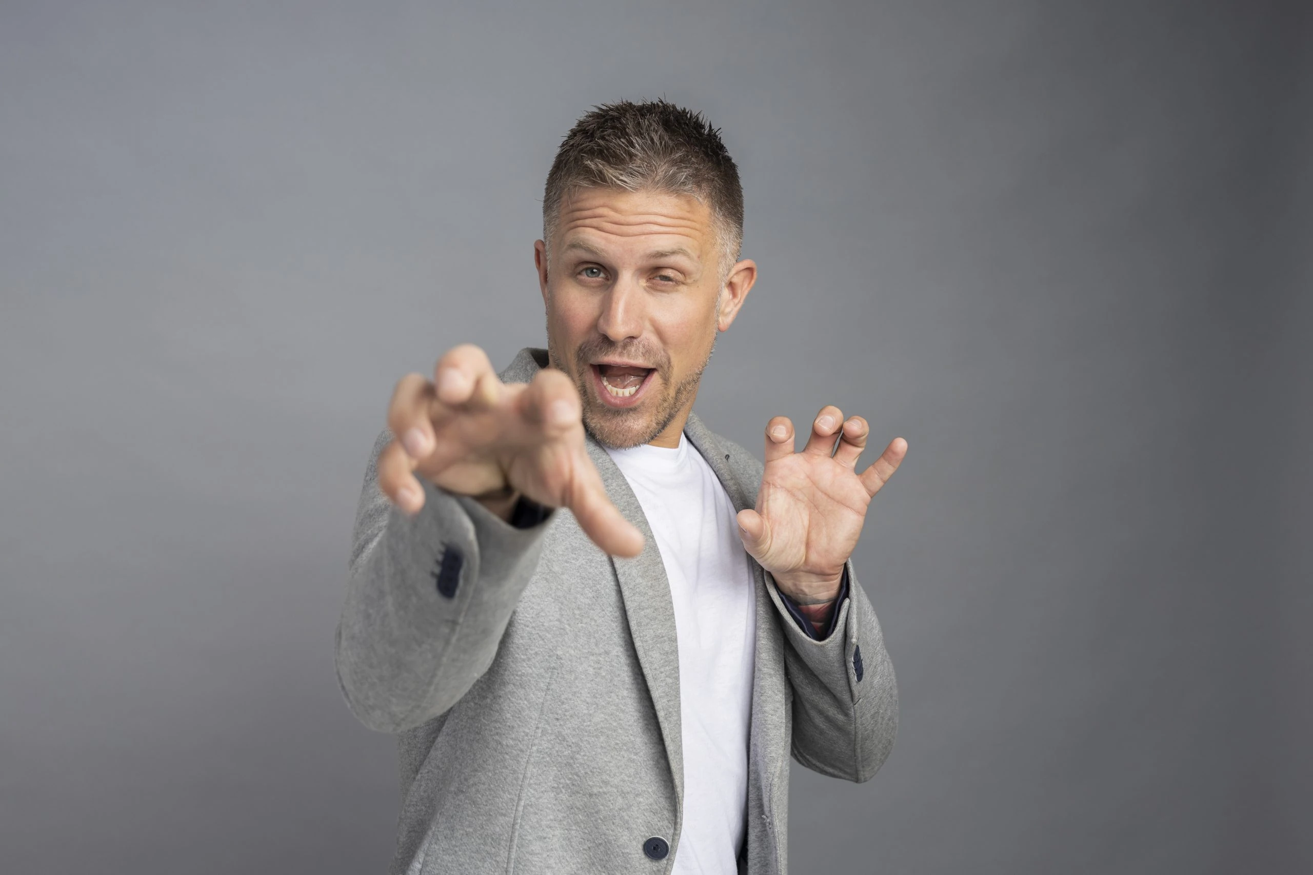 Man in a gray blazer and white shirt playfully gestures with hands against a gray background, embodying the sophistication and charisma often seen in advanced property management professionals.