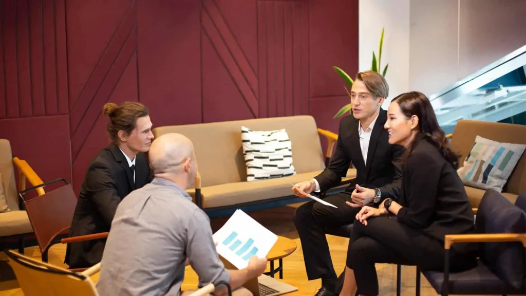 Four people in business attire are seated in a modern office setting, engaged in a discussion. One holds a paper with graphs, likely analyzing trends for property management companies.