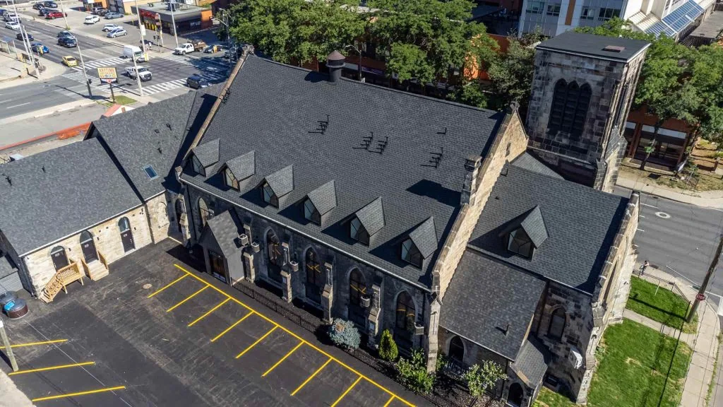 Aerial view of a large stone church with a steep roof, surrounded by an advanced property management complex, including rental spaces. Adjacent parking lot and nearby street intersection complete the Hamilton city's landscape.