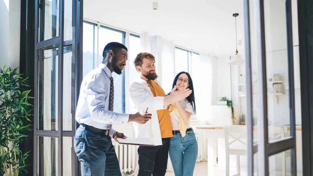 In an airy modern room, a representative from a prominent property management company in Hamilton gestures animatedly while explaining something to two attentive listeners. Lush plants and expansive windows create a serene backdrop, enhancing the engaging conversation.