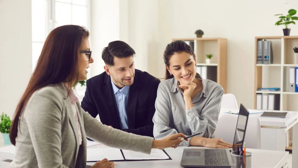 Three tenants sit at a desk in an office setting, discussing information on a laptop. One is pointing at the screen, highlighting important details. Shelves and plants add a touch of greenery to the background, creating an inviting atmosphere for their discussion.