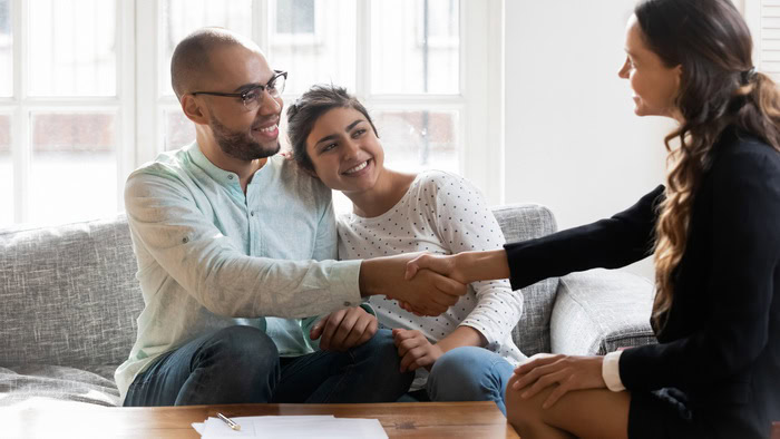 A couple sits on a couch smiling as they shake hands with a woman in business attire across a table with papers, finalizing their decision to enlist her expert property management services.