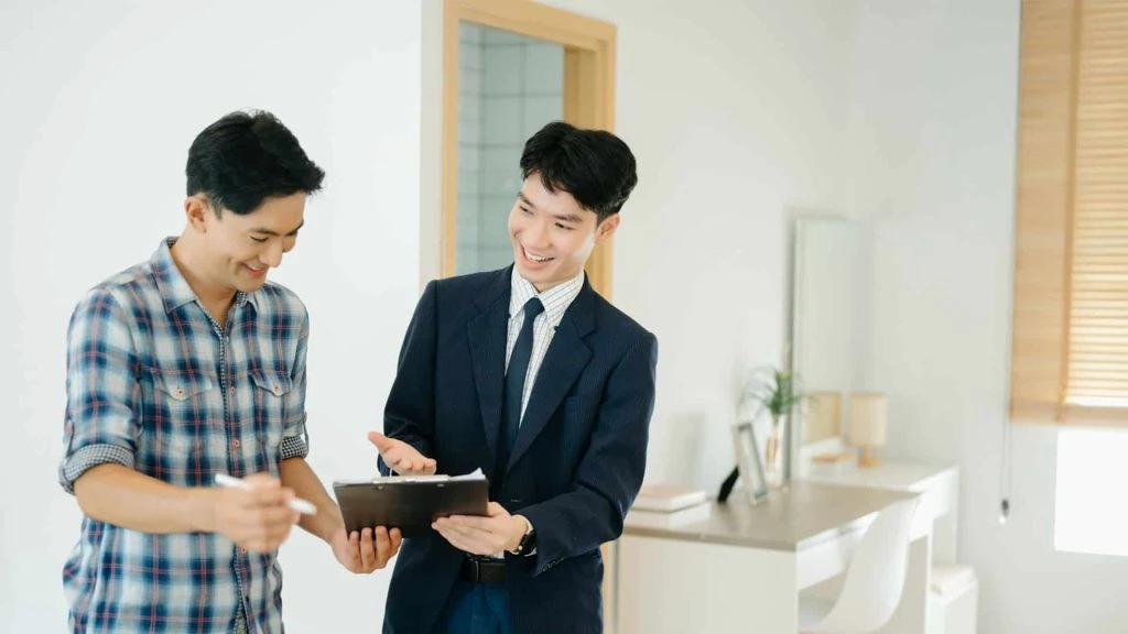 Two people stand indoors. One, in a suit and brandishing a clipboard, represents Hamilton Rentals. He appears to explain something engagingly to the other person, who is casually dressed and smiling with interest.