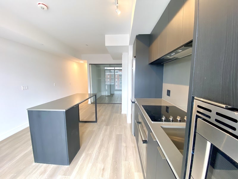 Modern kitchen courtesy of a rental company, featuring black cabinets, a built-in oven, and an induction cooktop. Light wood flooring complements a large kitchen island and a sliding door that leads to another room.