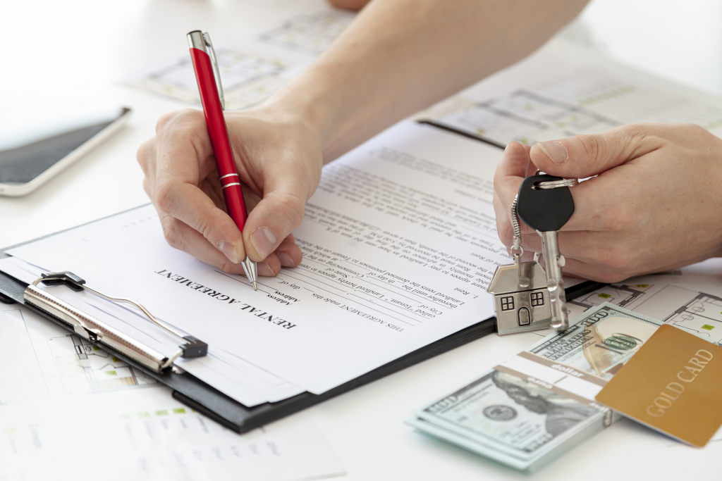 A person signs an Ontario Landlord and Tenant rental agreement on a clipboard while holding house keys, a small house keychain, and a stack of money with a gold credit card.