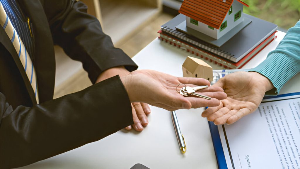 A person in a suit hands over house keys to another person across a desk with Ontario Landlord and Tenant documents, a pen, and model houses.