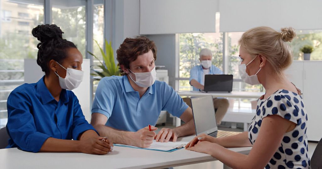 Three people wearing face masks sit at a table discussing documents related to a background check for tenants, while another person works on a laptop in the background.