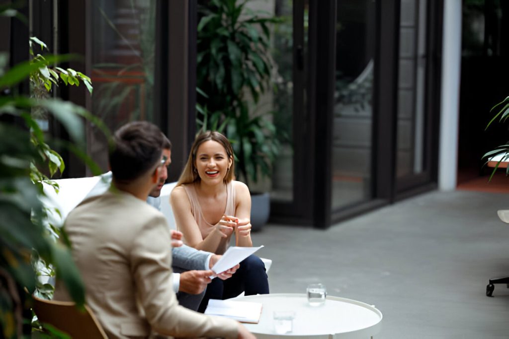 Two people sit at a round table with documents discussing a background check for tenants, glasses of water between them in a bright, modern indoor space with large windows and plants.