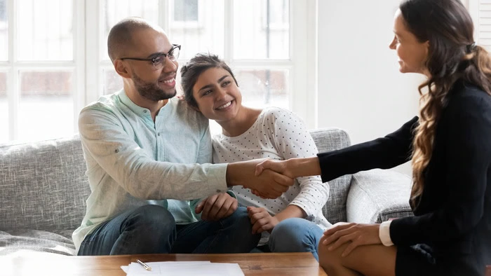 A couple sits on a couch smiling as the woman shakes hands with a professional across a table, discussing their Month-to-Month Lease Ontario agreement.