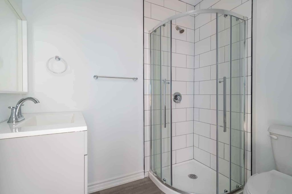 A modern bathroom with a corner glass shower, white vanity with chrome faucet, towel bar, and toilet, featuring white tiled walls and light wood flooring—perfectly maintained and ready to Inspect Property.