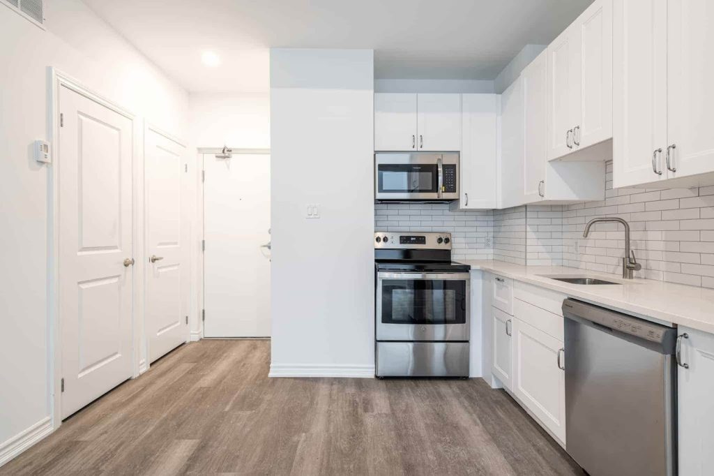 Modern kitchen with white cabinets, stainless steel appliances, subway tile backsplash, and wood-look flooring. Two closed doors are visible on the left side—ideal to inspect property features firsthand.
