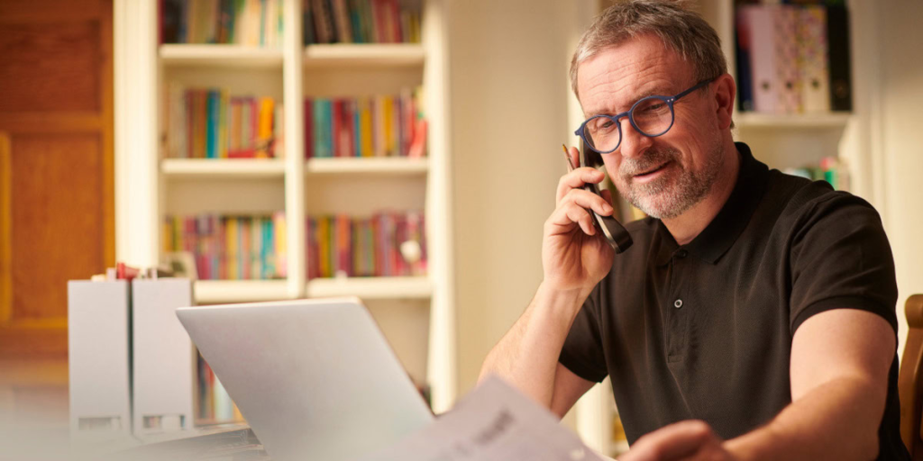 A man wearing glasses sits at a desk, talking on the phone and looking at papers—perhaps calling to identify a property owner—with a laptop and bookshelves in the background.