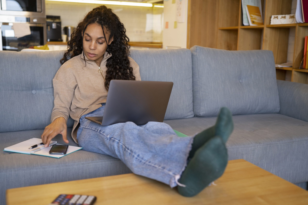A person sits on a sofa with a laptop on their lap, writing in a notebook while reaching for a smartphone—perhaps researching how to identify a property owner.