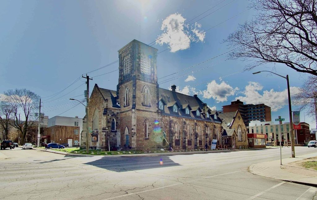 A stone church with a square tower sits at a street corner on a sunny day, surrounded by trees, modern buildings, and parked cars—an inviting spot to inspect property in this charming neighborhood.