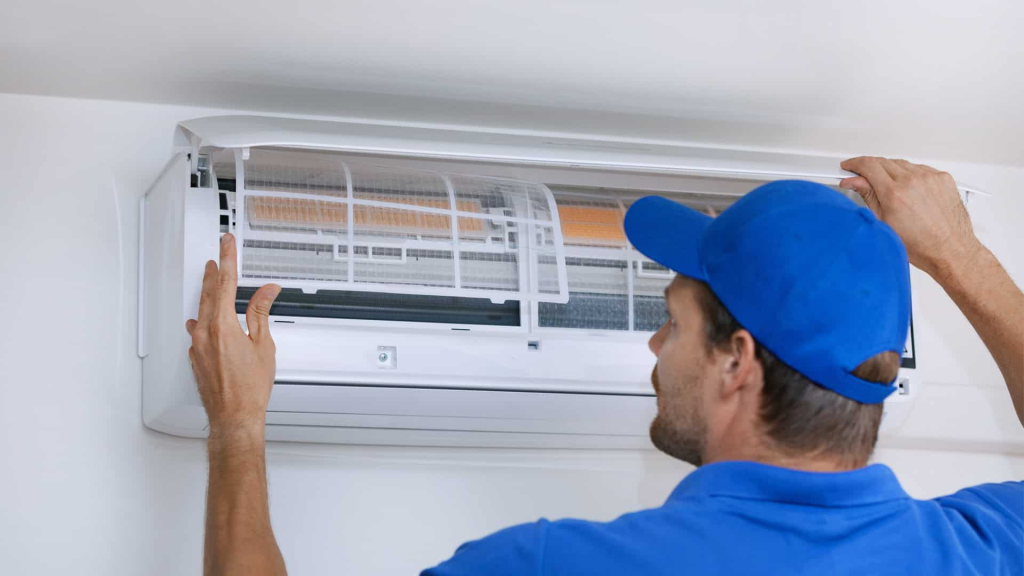 A technician in a blue uniform lifts the cover of a wall-mounted air conditioning unit for inspection or maintenance as part of property maintenance in Hamilton.
