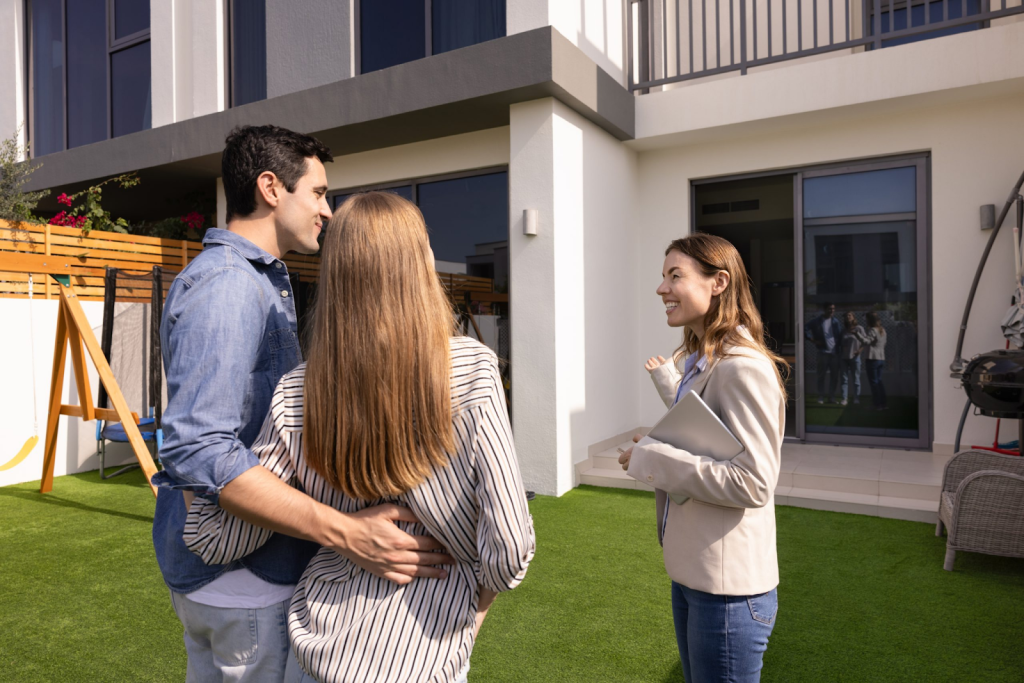 A couple stands outside a modern house, listening to a woman holding a folder, likely a real estate agent, discuss the details of renting with pets during their property viewing.